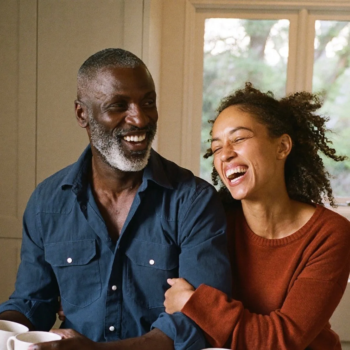 A man and his wife laughing together over coffee at their kitchen table, present for his marriage after retiring from alcohol
